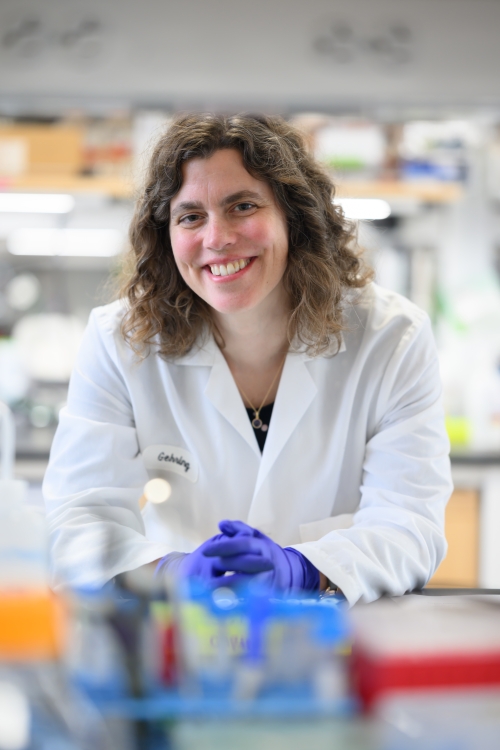 Mary Gehring, in a lab coat, smiles at the camera from across a lab bench