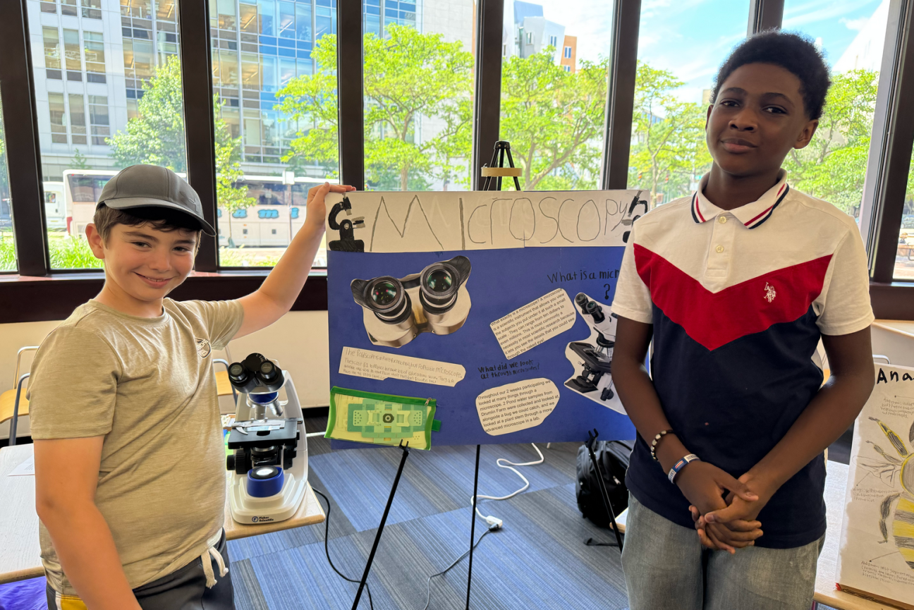 Two kids stand, one to either side of a poster on "Microscopy"