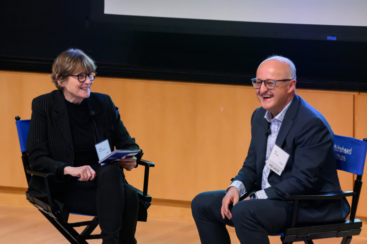 Two people sit in chairs facing partly towards each other and party towards an audience offscreen. Left, Ruth, has interview cards and looks at Uli, right. Both are smiling.