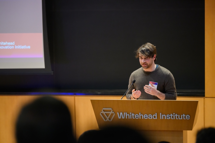 A man speaks from behind a podium, with a corner of the slidehshow visible to the side