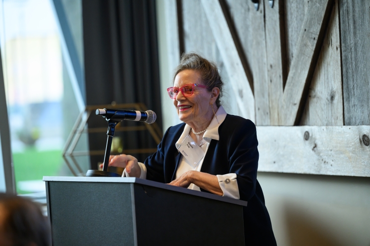 A woman speaks at a podium in front of a wooden barn door, with grass visible outside