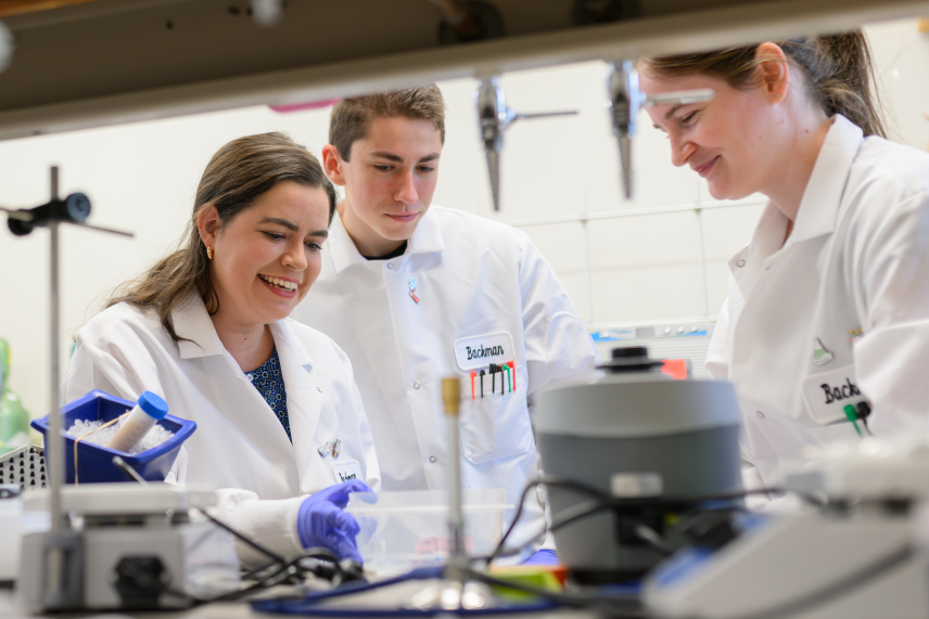 From across a lab bench, we see three researchers in white coats looking at something on the bench