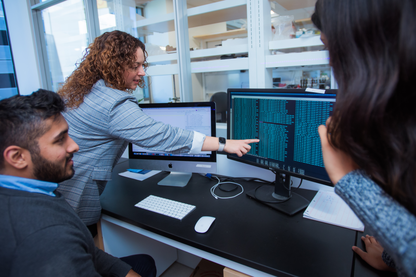 Olivia Corradin points to a line on a computer screen with many lines of text and data, while two lab members look on